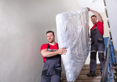 Two,Young,Male,Movers,In,Uniform,Carrying,The,Wrapped,Mattress